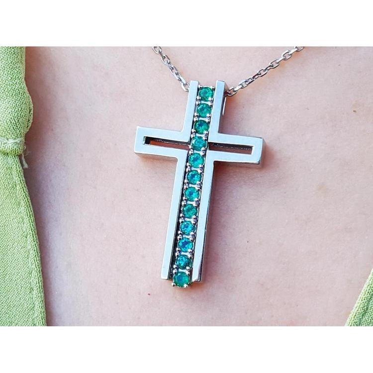 A white gold cross pendant with round cut natural emeralds on a chain, worn on a person's neck.