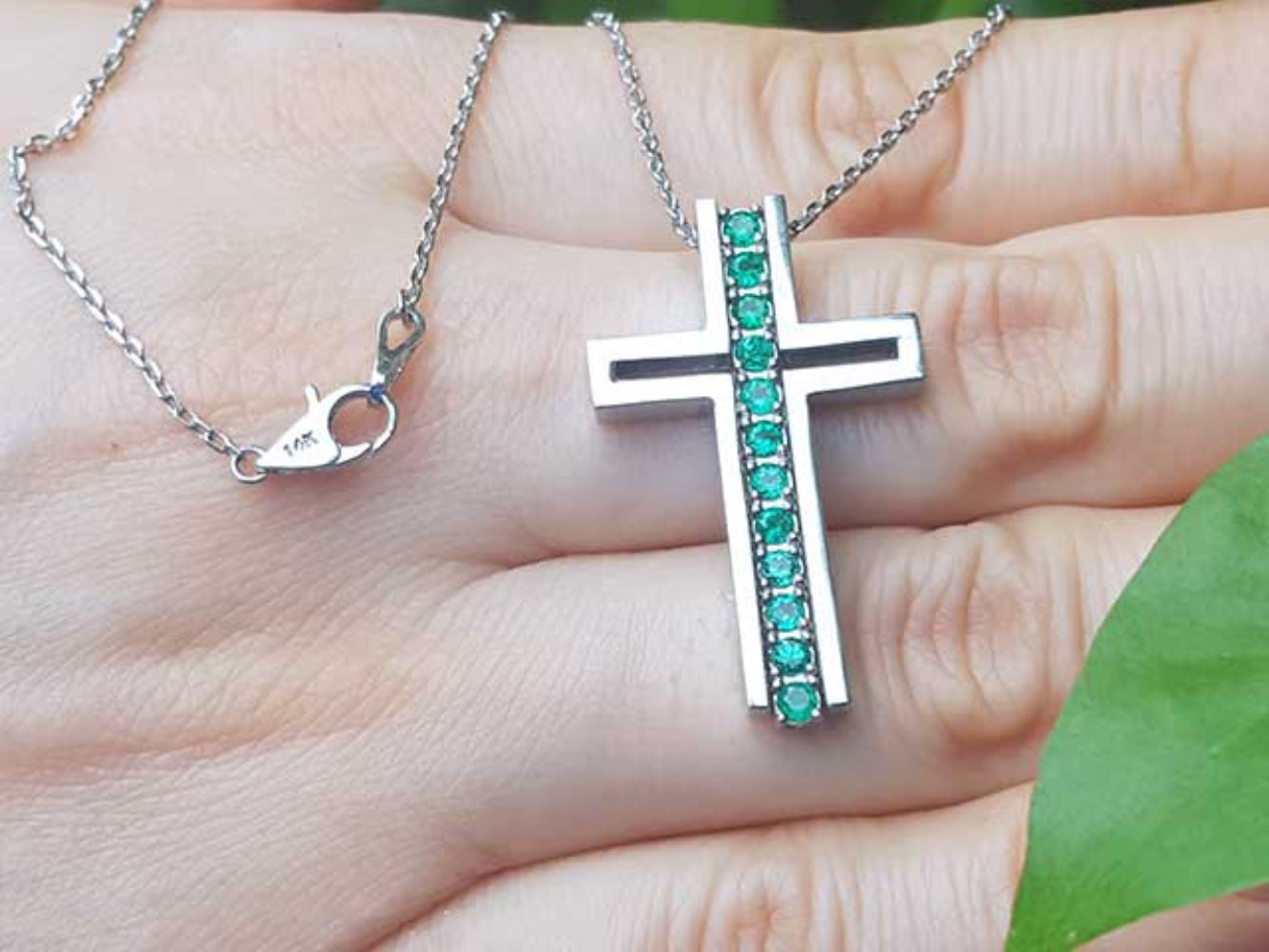 White gold cross pendant necklace with round emeralds held in a hand.
