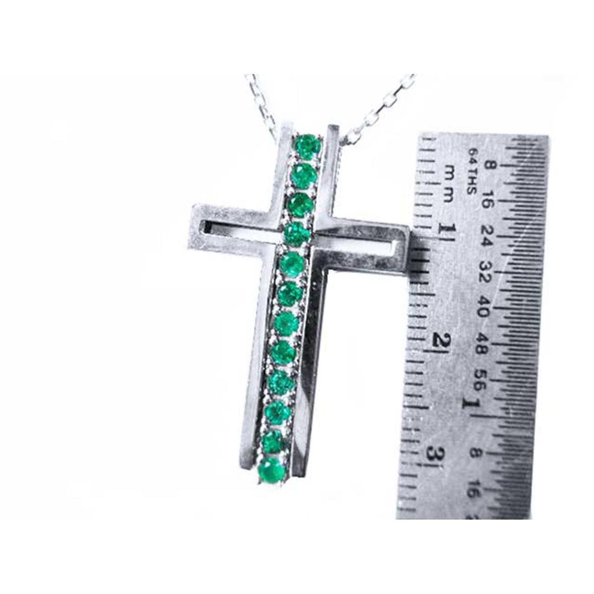 A white gold cross pendant with natural emeralds next to a ruler for scale on a white background.