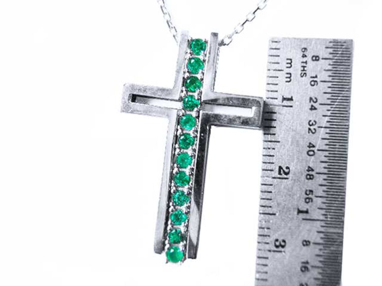 A white gold cross pendant with natural emeralds next to a ruler for scale on a white background.
