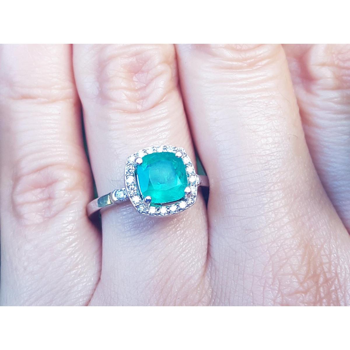 A close-up of a cushion cut emerald surrounded by a halo diamond in white gold on a person's hand.