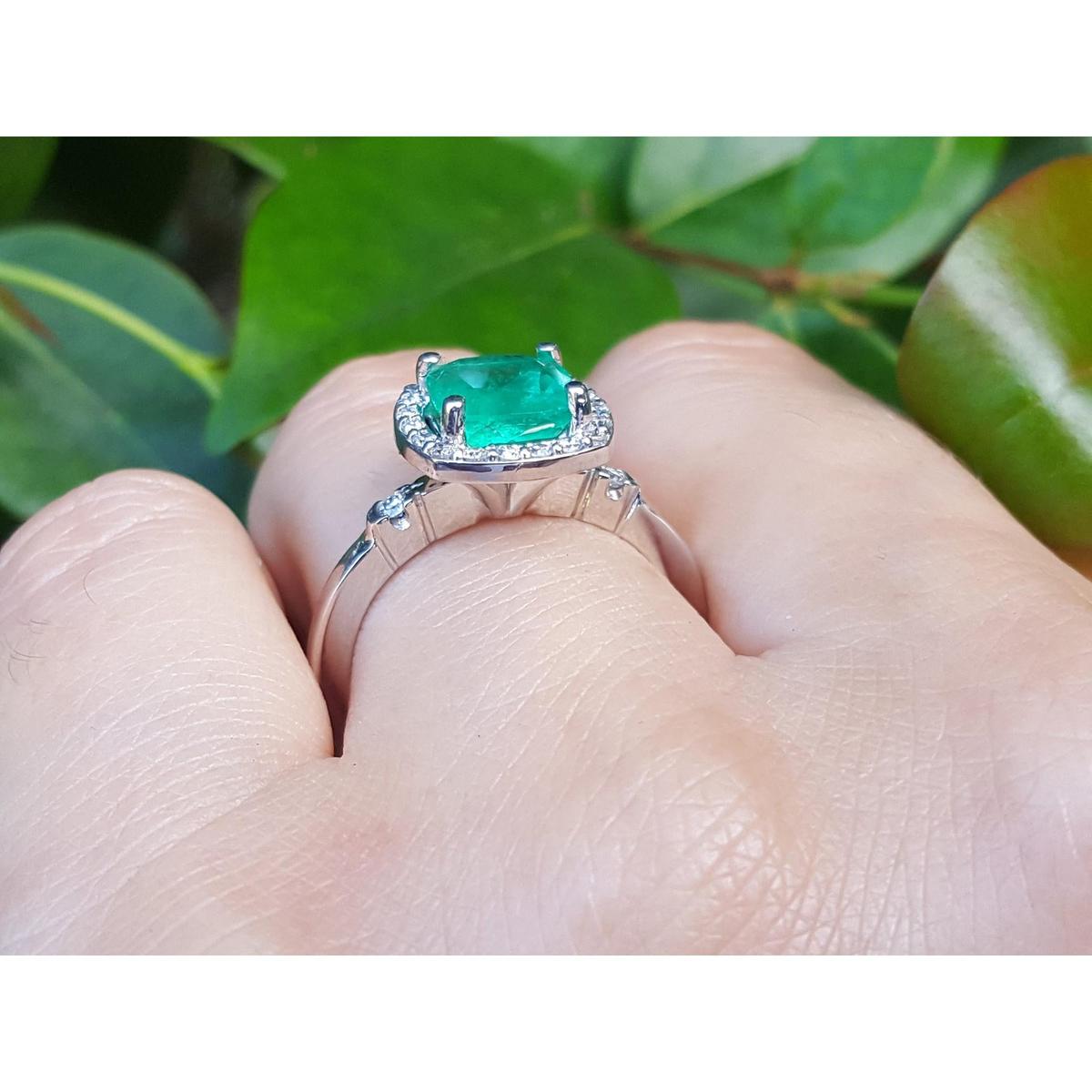 On top of a finger wearing a white gold cushion cut emerald ring on a leafy background.