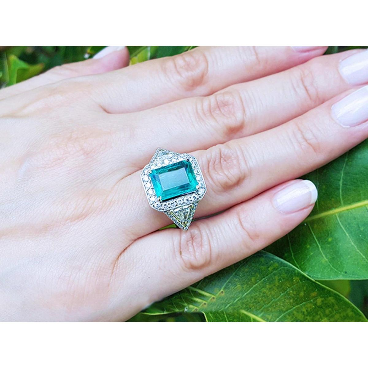 Hand wearing an emerald ring with side trillion cut diamonds against a leafy background.