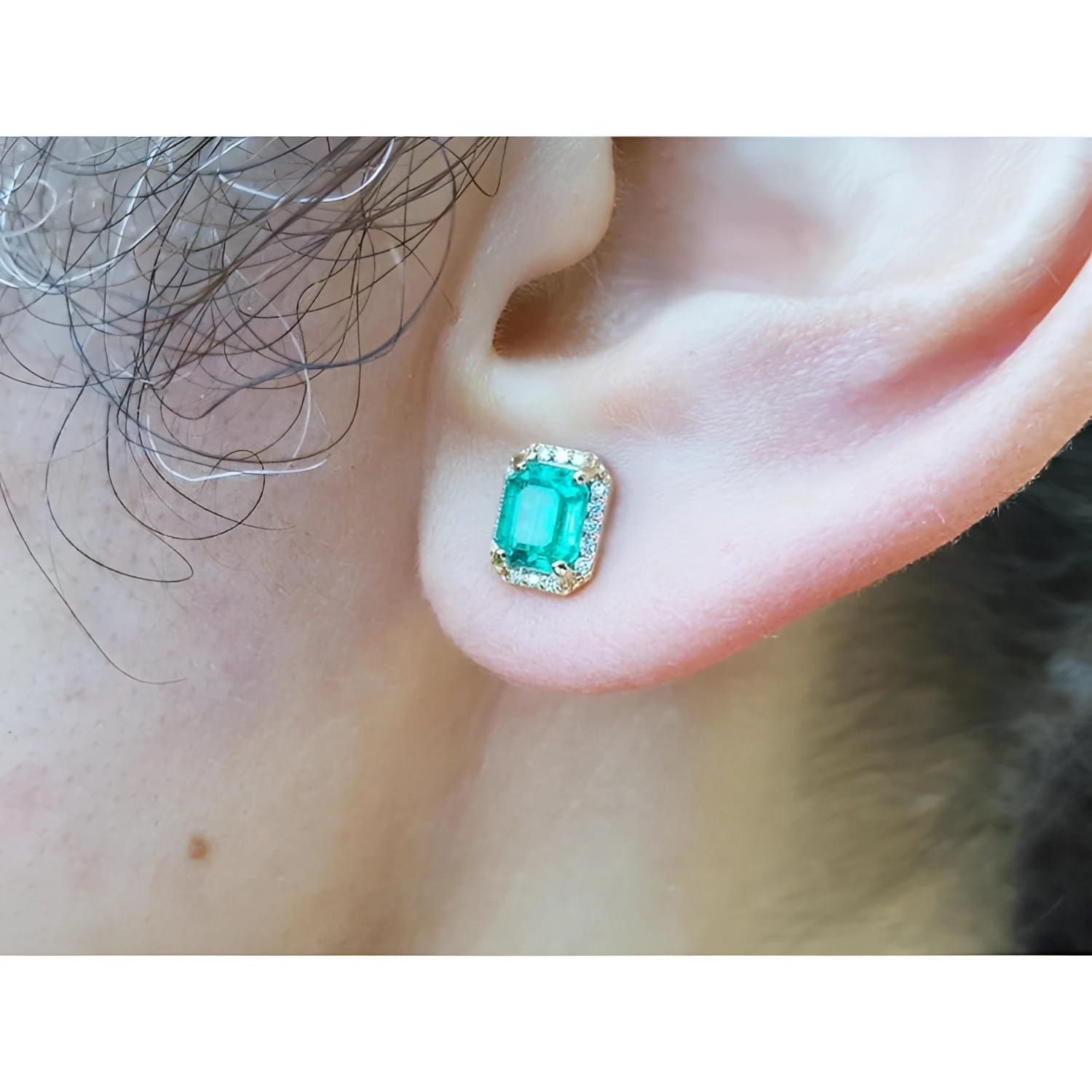 An ear wearing a natural emerald stud earring with halo diamond in 14k gold.