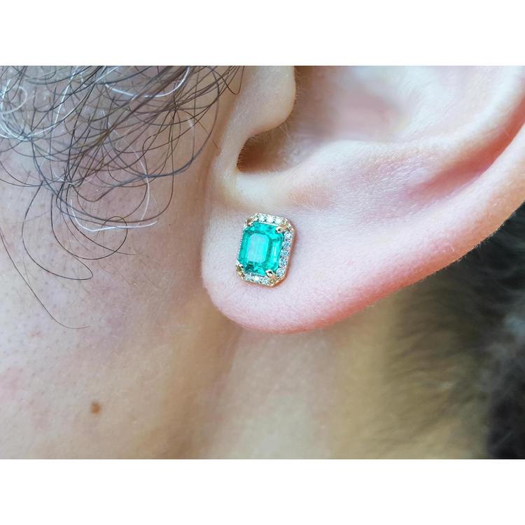 An ear wearing a natural emerald stud earring with halo diamond in 14k gold.