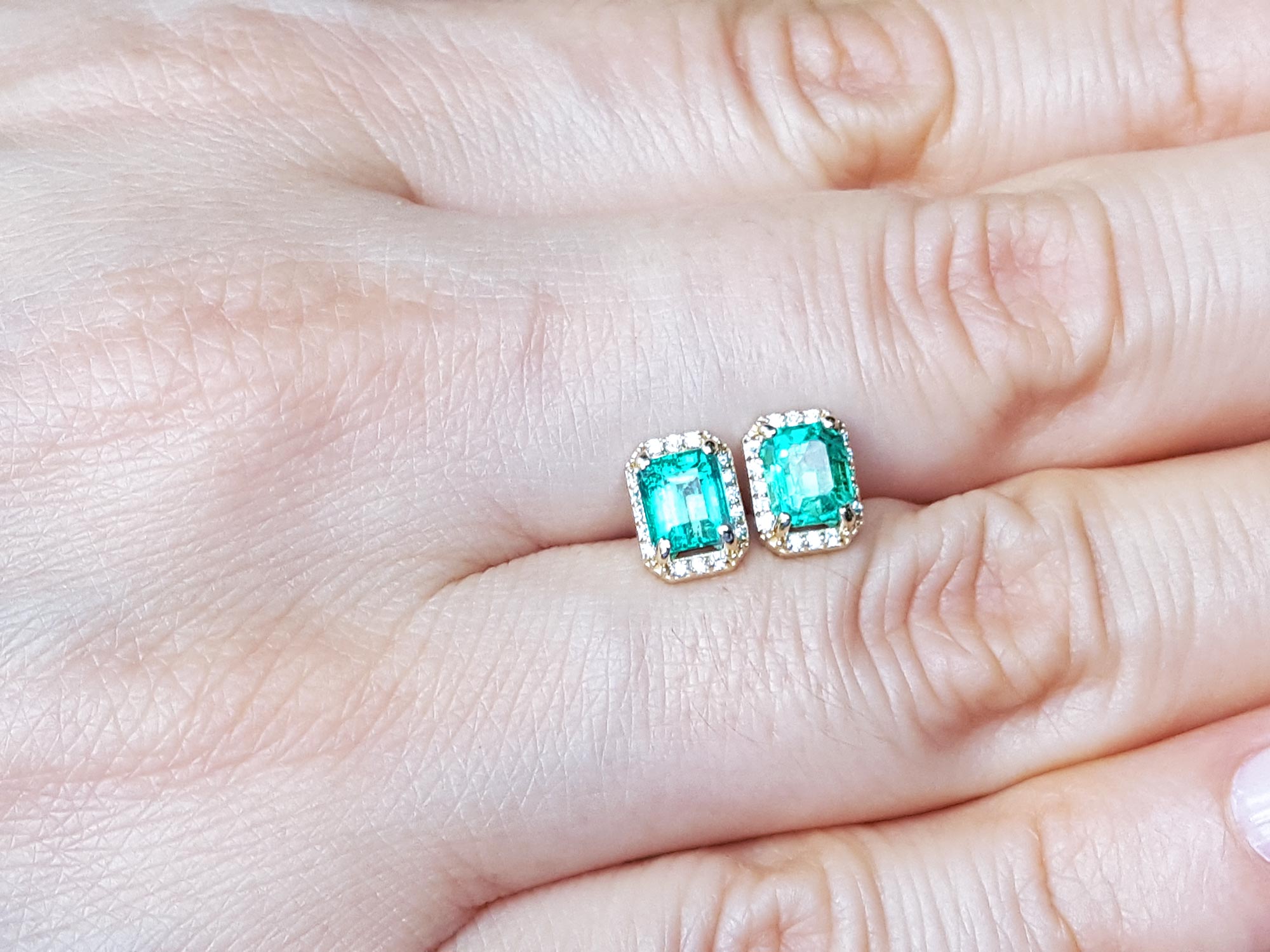 Emerald green gemstone earrings held between fingers on a light skin background.