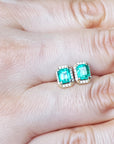 Emerald green gemstone earrings held between fingers on a light skin background.