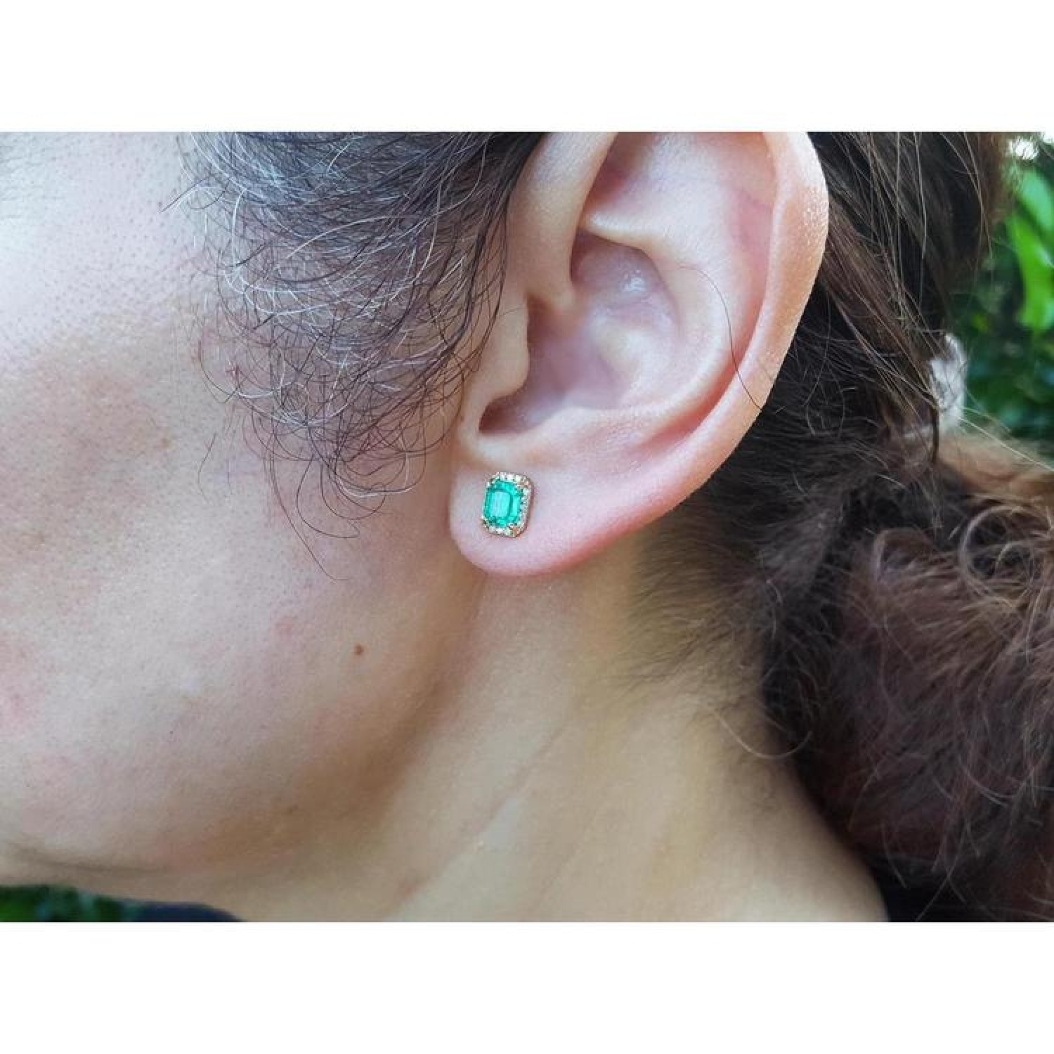 Rectangular Colombian emerald halo stud earrings in 14k yellow gold, worn on an ear.