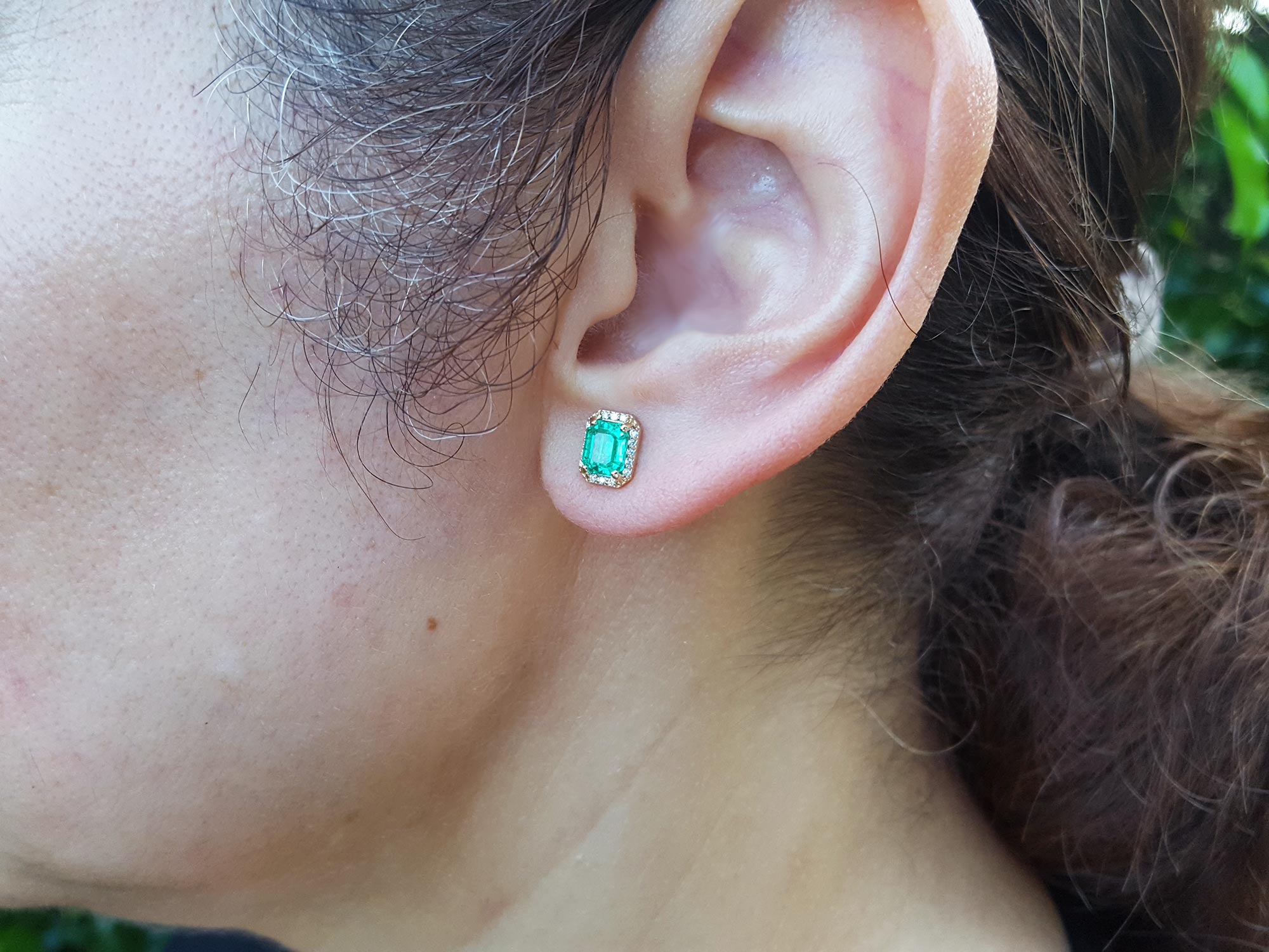 Close-up of a woman&#39;s ear wearing a yellow gold emerald stud earring with blurred background.
