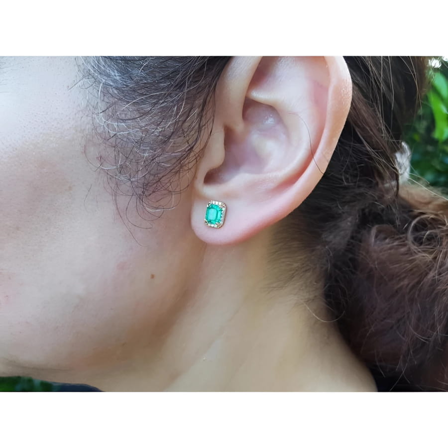 Rectangular Colombian emerald halo stud earrings in 14k yellow gold, worn on an ear.