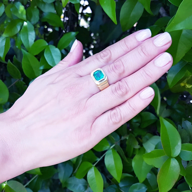 Hand wearing a 18k yellow gold emerald ring for men against a green leafy background.