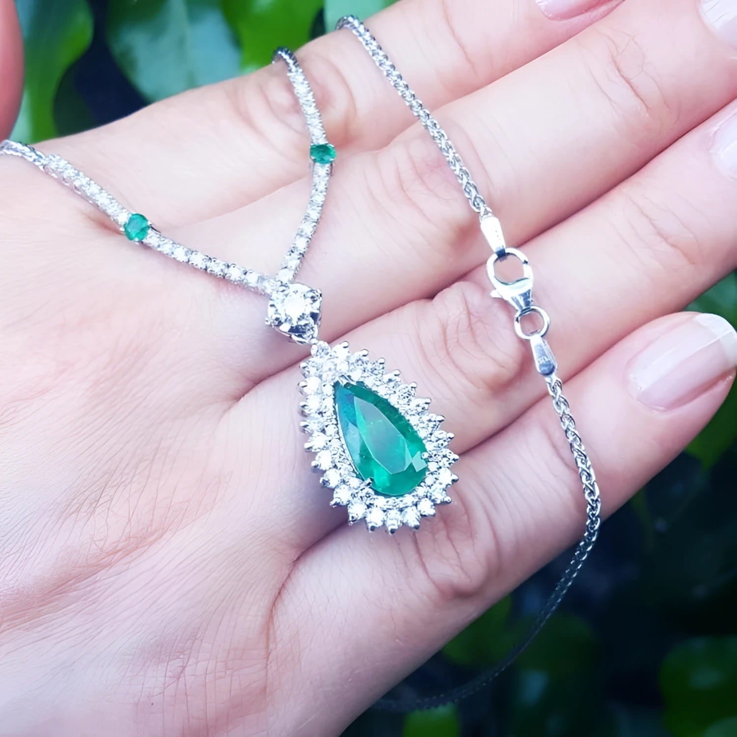 Pear cut emerald necklace with double diamond halo on 18K white gold chain, held in hand.