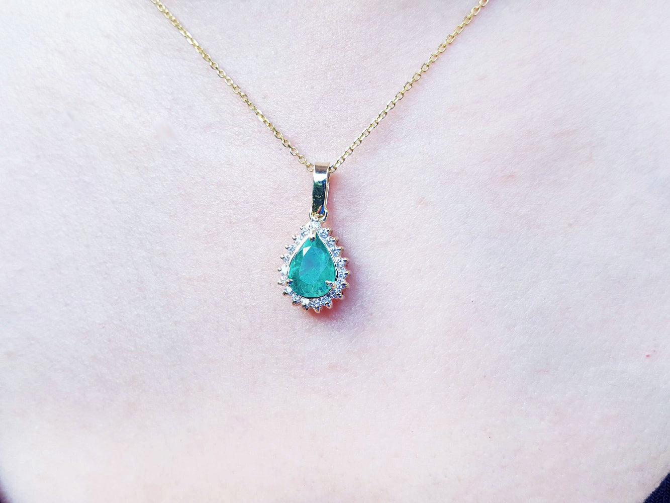 Teardrop-shaped colombian emerald pendant on a gold chain against a light pink skin background.