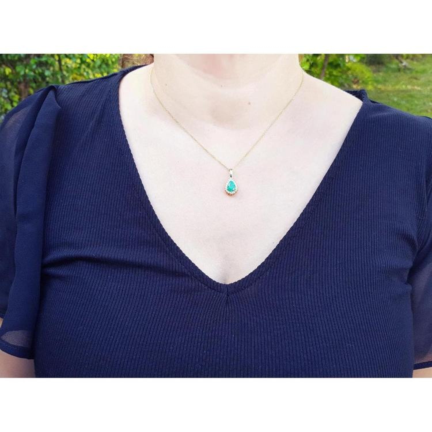 Pear-shaped Colombian emerald halo diamond pendant on a gold chain, worn on a navy top.
