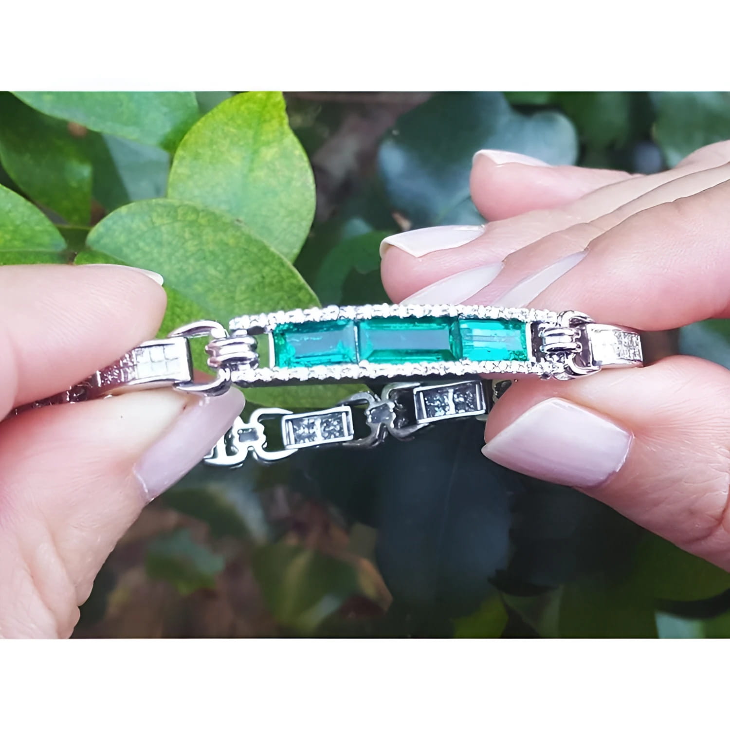 Emerald and princess cut diamond bracelet held against green leaves.