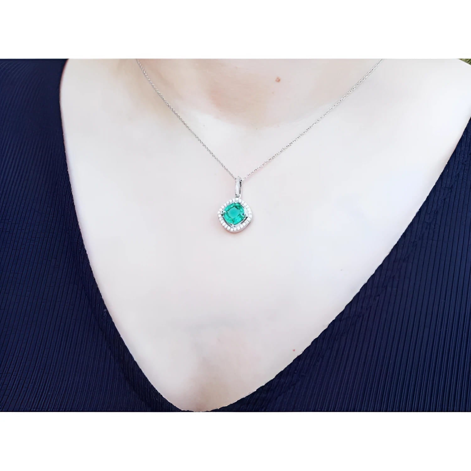 Cushion-cut Colombian emerald pendant with diamond halo on a white gold chain, worn on a dark top.