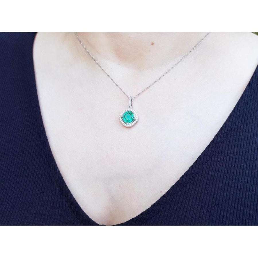 Cushion-cut Colombian emerald pendant with diamond halo on a white gold chain, worn on a dark top.