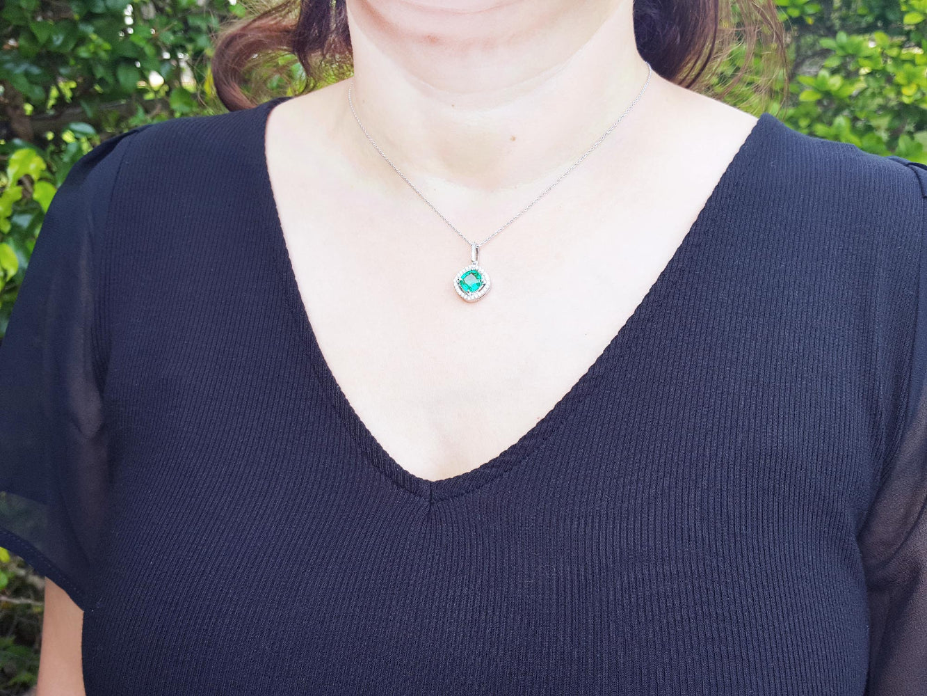 A woman wearing a black top with a necklace featuring a natural emerald pendant against a blurred green background.