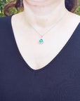 A woman wearing a black top with a necklace featuring a natural emerald pendant against a blurred green background.