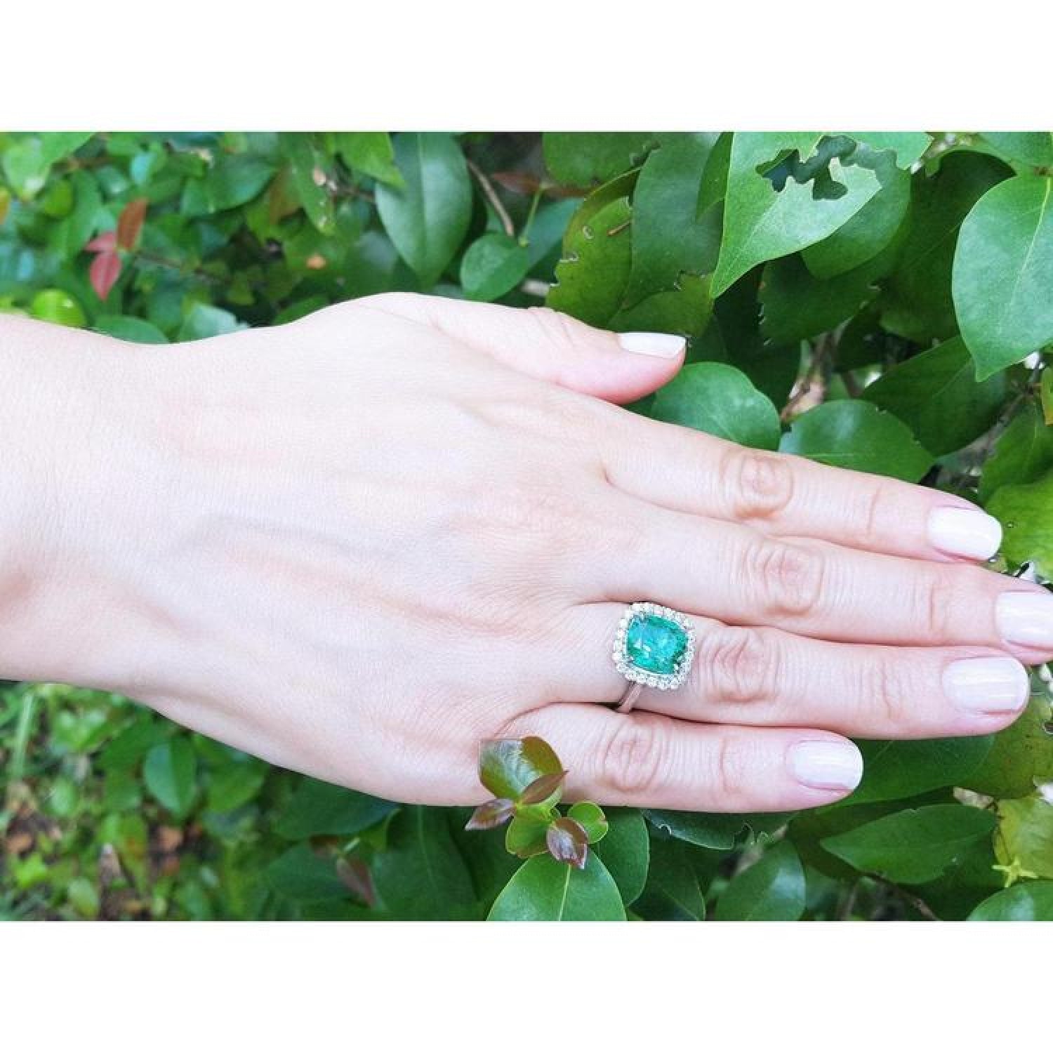 Cushion cut Colombian emerald engagement ring with diamond halo in 18K white gold on a hand.