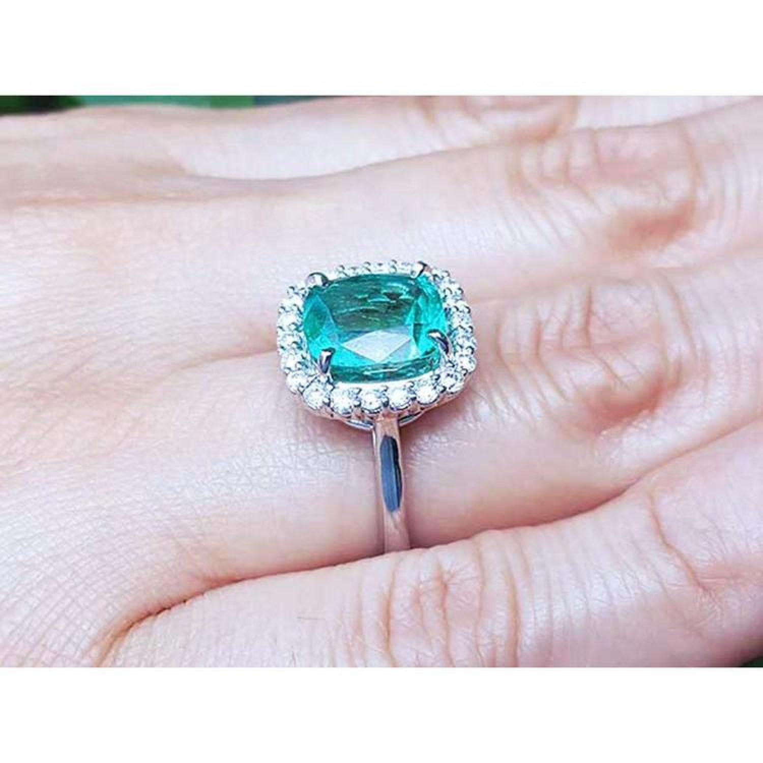 Cushion cut Colombian emerald ring with diamond halo in 18K white gold on a hand.
