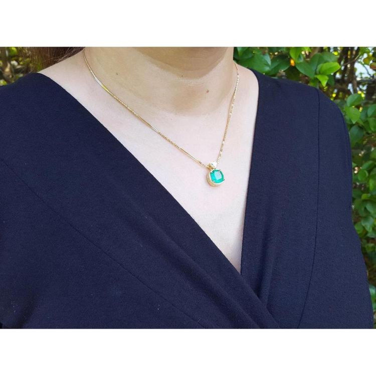 Woman wearing a cushion cut emerald pendant necklace with a small diamond accent on an 18K yellow gold chain.