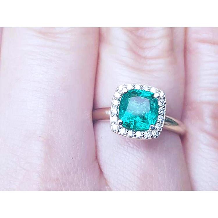 A women's cushion cut emerald ring with halo diamond in 14k yellow gold on a finger.