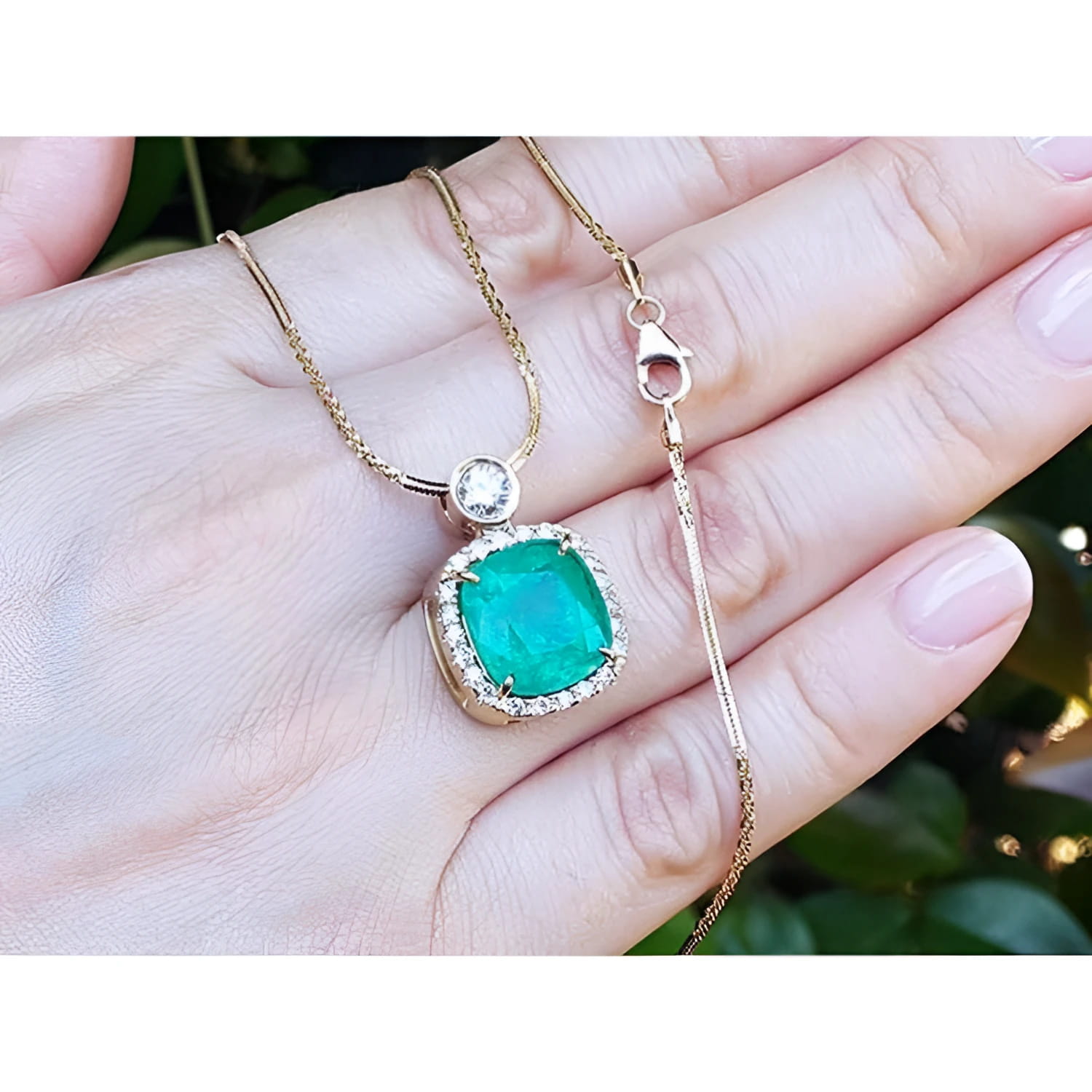 Cushion cut emerald necklace with halo diamonds on an 18K yellow gold chain, held in a hand.