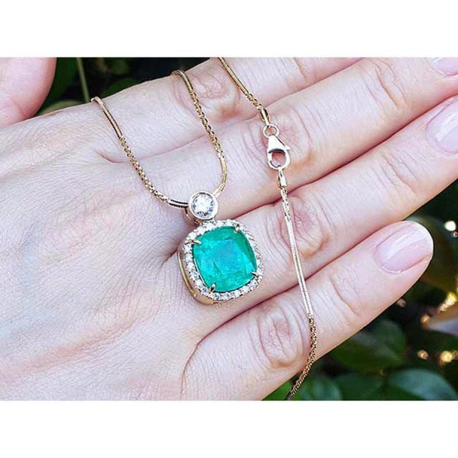 Cushion cut emerald necklace with halo diamonds on an 18K yellow gold chain, held in a hand.