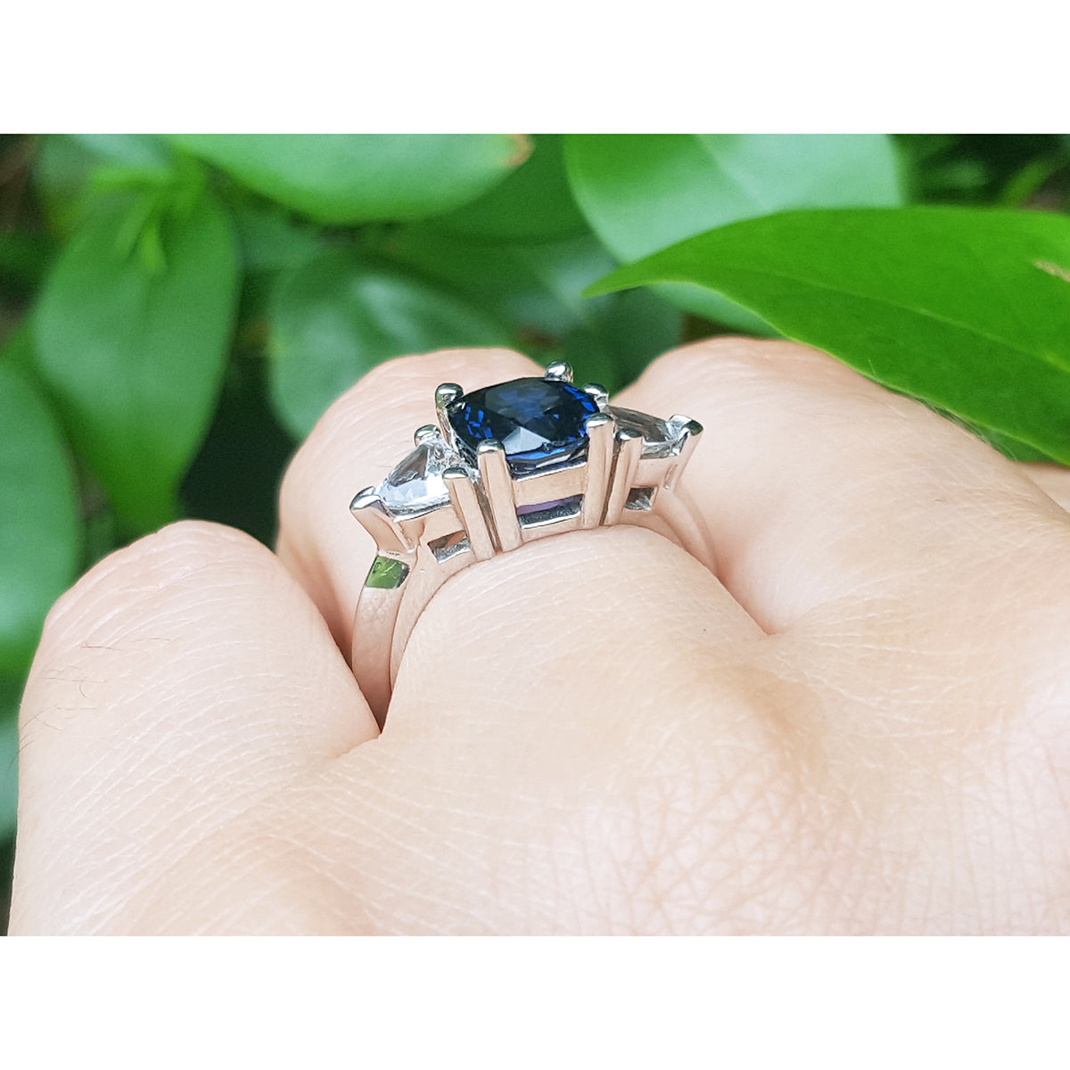 White gold ring with a blue sapphire cushion cut held in a hand against a green leafy background