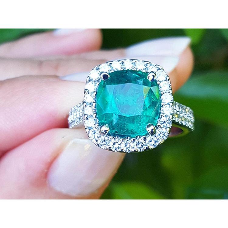 Cushion cut natural colombian emerald ring with halo diamond made of 14k white gold held between fingers.