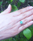Hand wearing a cushion cut emerald ring with a leafy background.