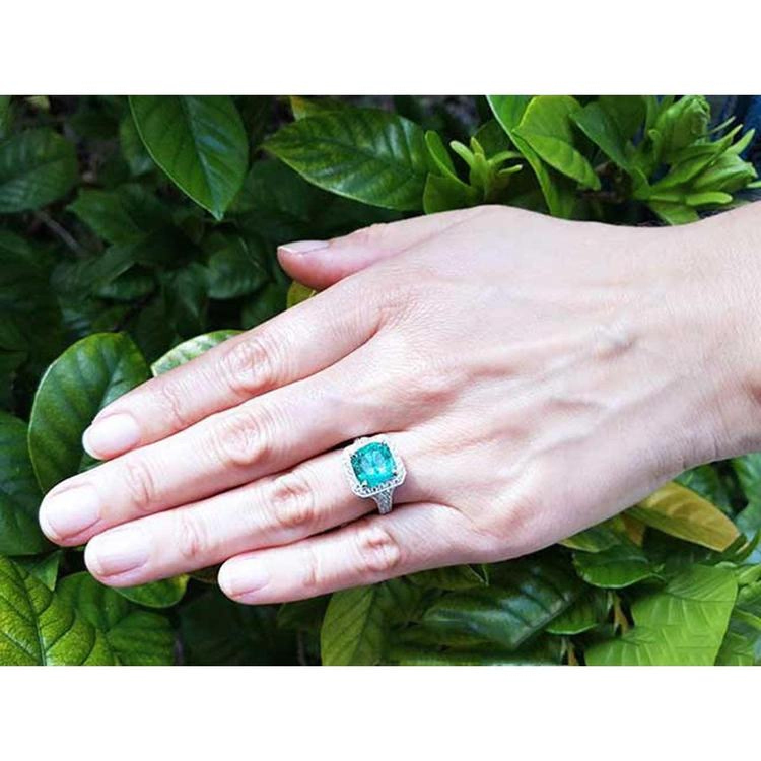 Cushion cut emerald engagement ring with diamond halo in 18K white gold on a hand.