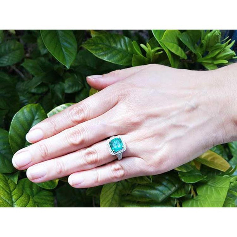 Cushion cut emerald engagement ring with diamond halo in 18K white gold on a hand.
