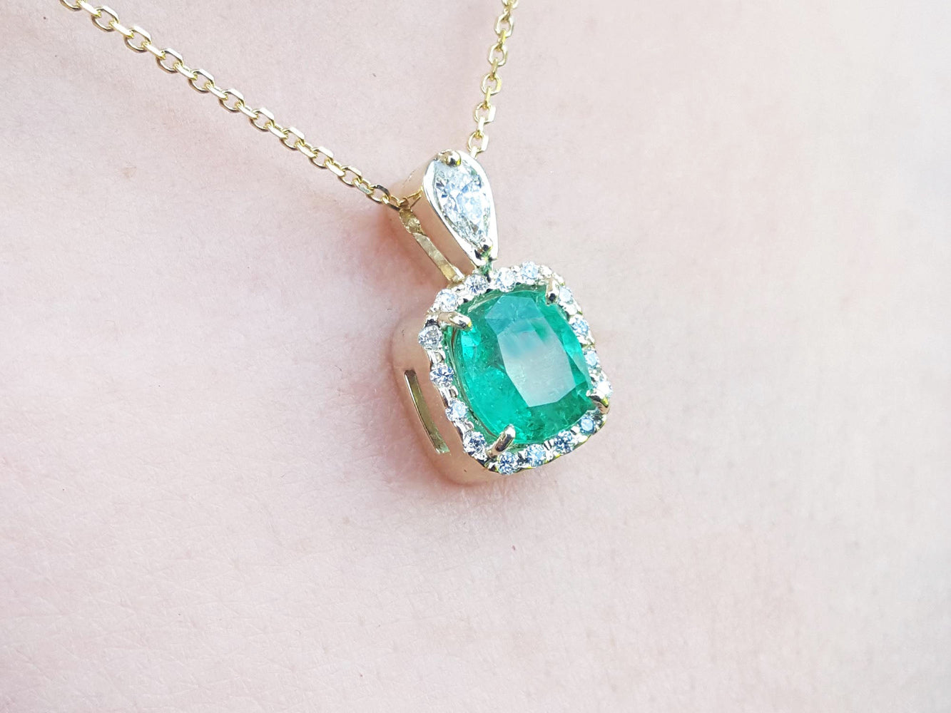 A natural emerald gemstone pendant on a gold chain against a light pink background.