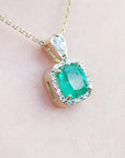 A natural emerald gemstone pendant on a gold chain against a light pink background.