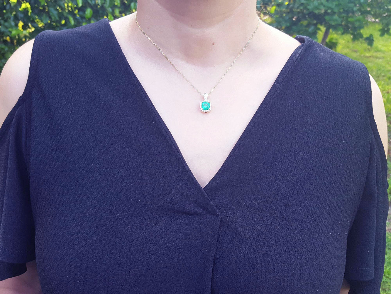 Navy blue dress with a cushion cut emerald pendant necklace on a woman&#39;s neck against a green outdoor background.