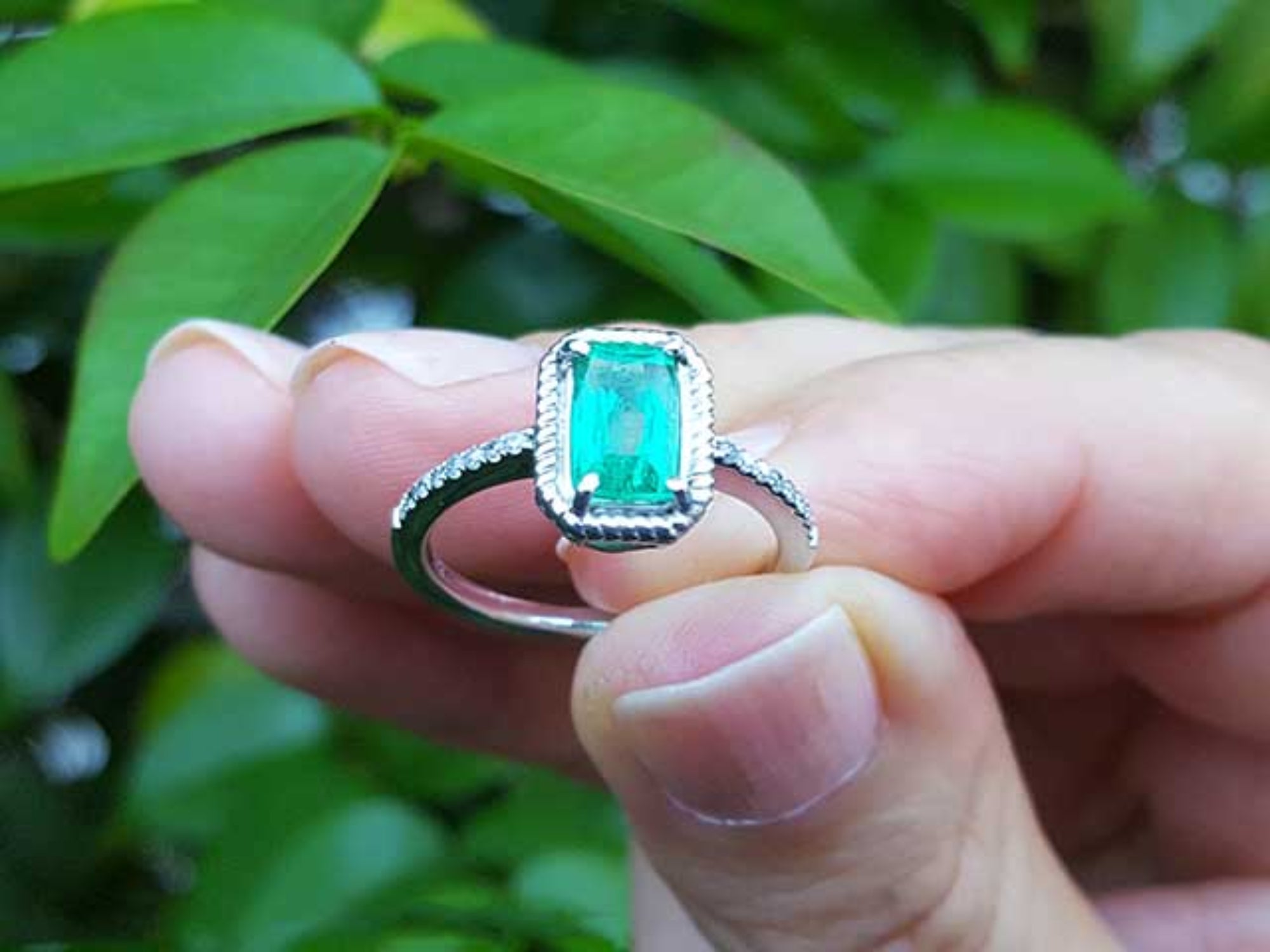 A natural emerald ring held between fingers against a blurred green leafy background.