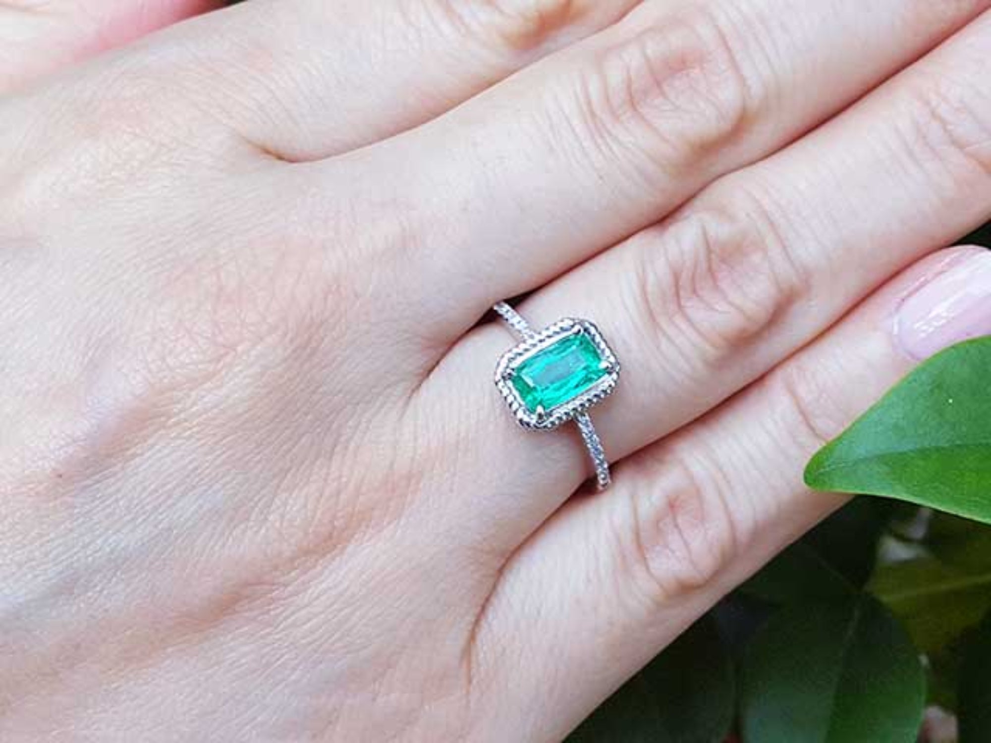 A cushion cut emerald ring on a hand with a green leaf background.