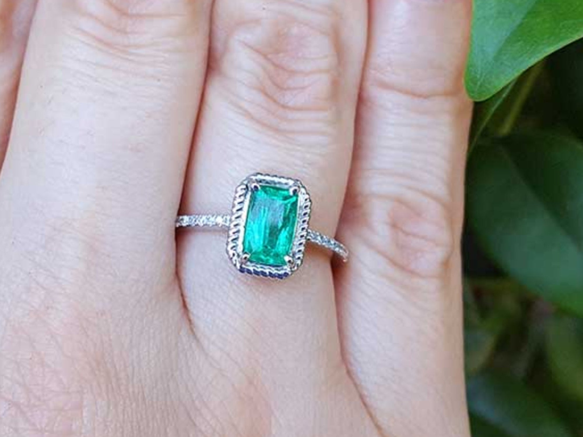 A cushion cut emerald ring on a finger with a blurred green leaf background.