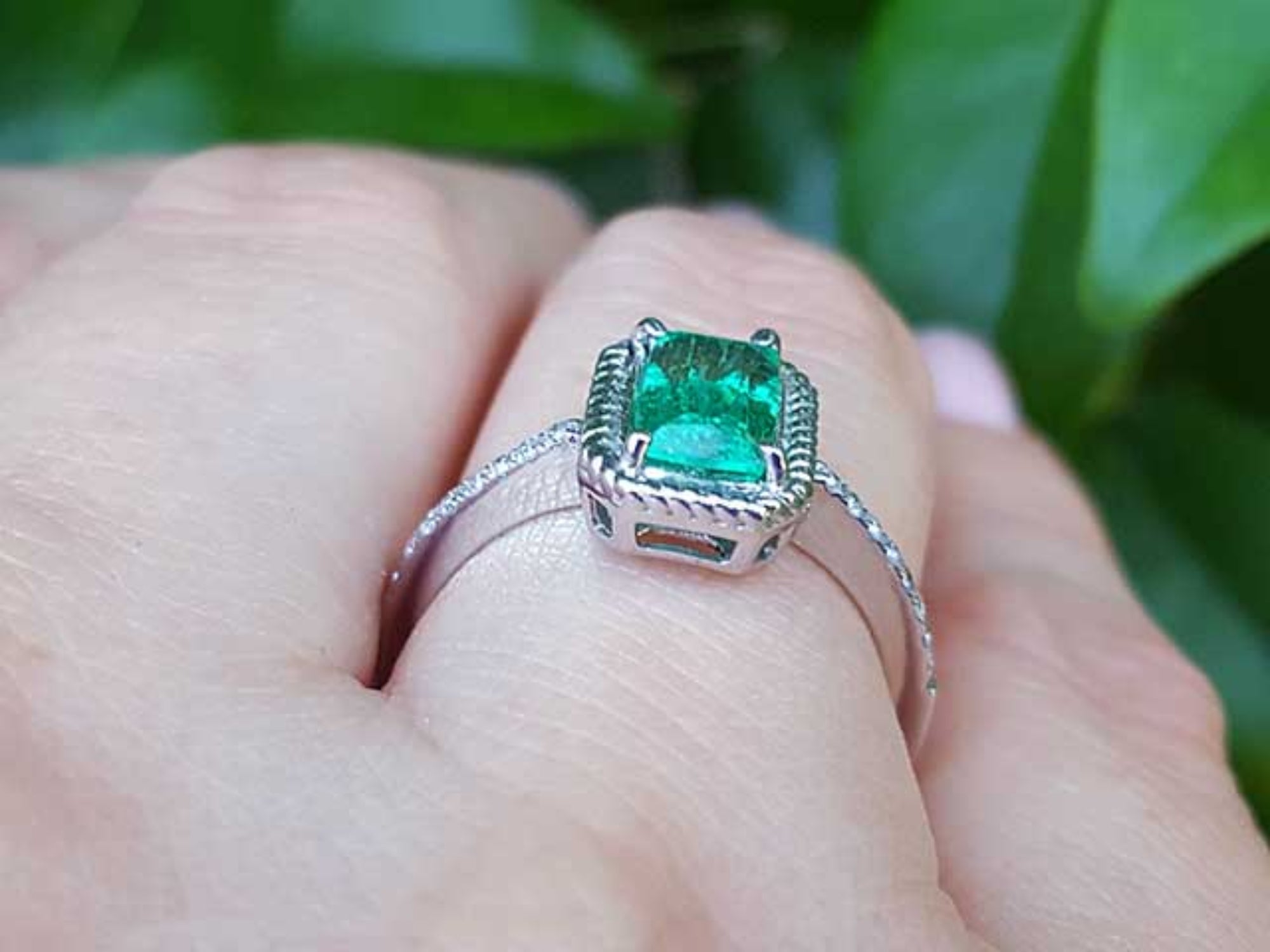 A white gold emerald ring on a hand against a green leafy background.