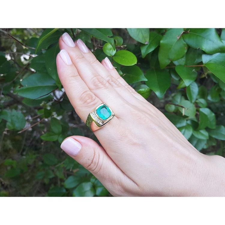 Hand wearing a gold men's ring with a cushion cut natural emerald against a green leafy background.