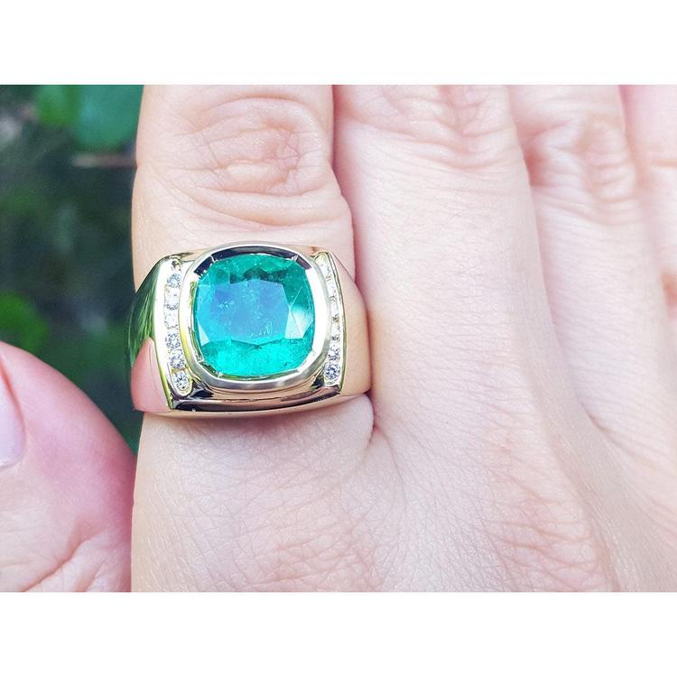 Gold ring for men with a cushion cut Muzo colombian emerald on a finger against a blurred natural background.
