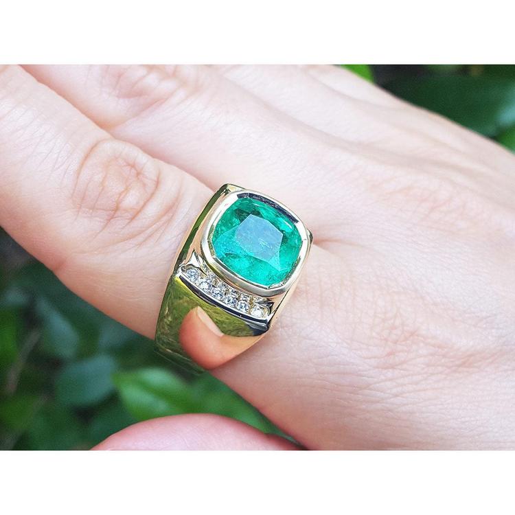 Gold ring for men with a natural emerald on a person's finger against a blurred green background.