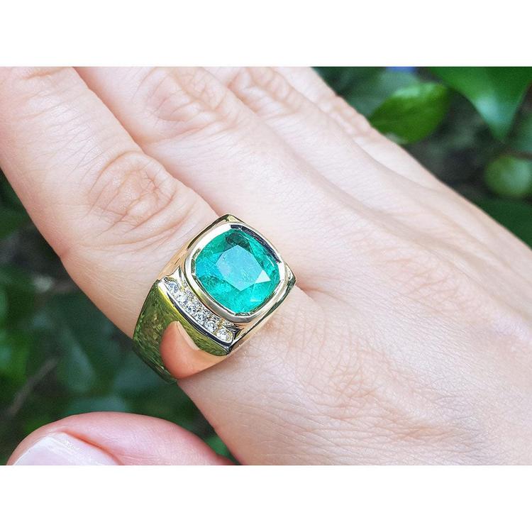 A 18k yellow gold mens emerald ring bezel set on a hand against a blurred green background.