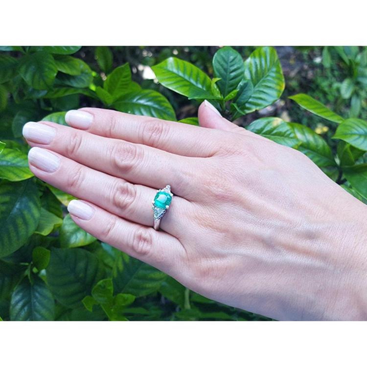 Cushion cut emerald ring with trillion side diamonds in 14K white gold on a hand.