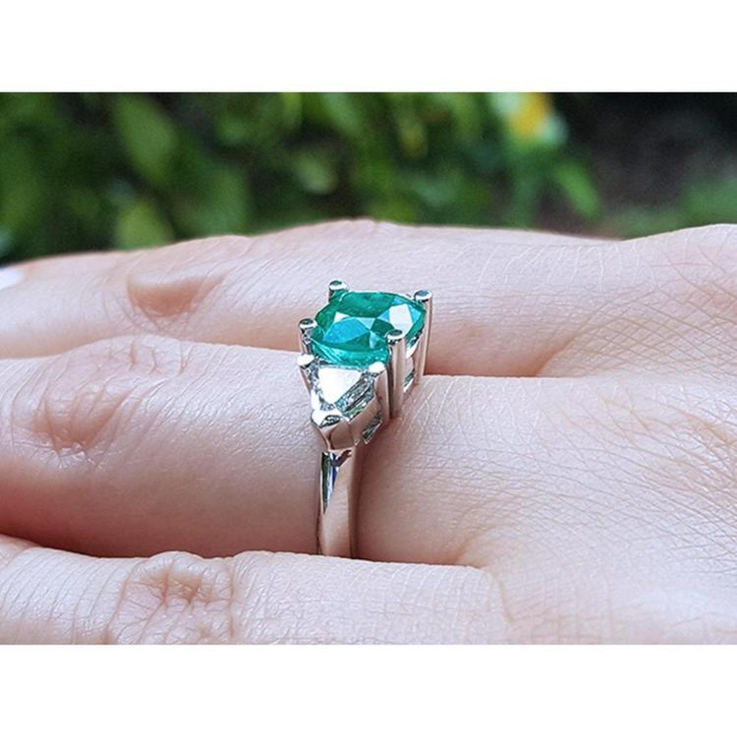 2.18 ct cushion cut emerald ring with trillion side diamonds in 14K white gold on a finger.