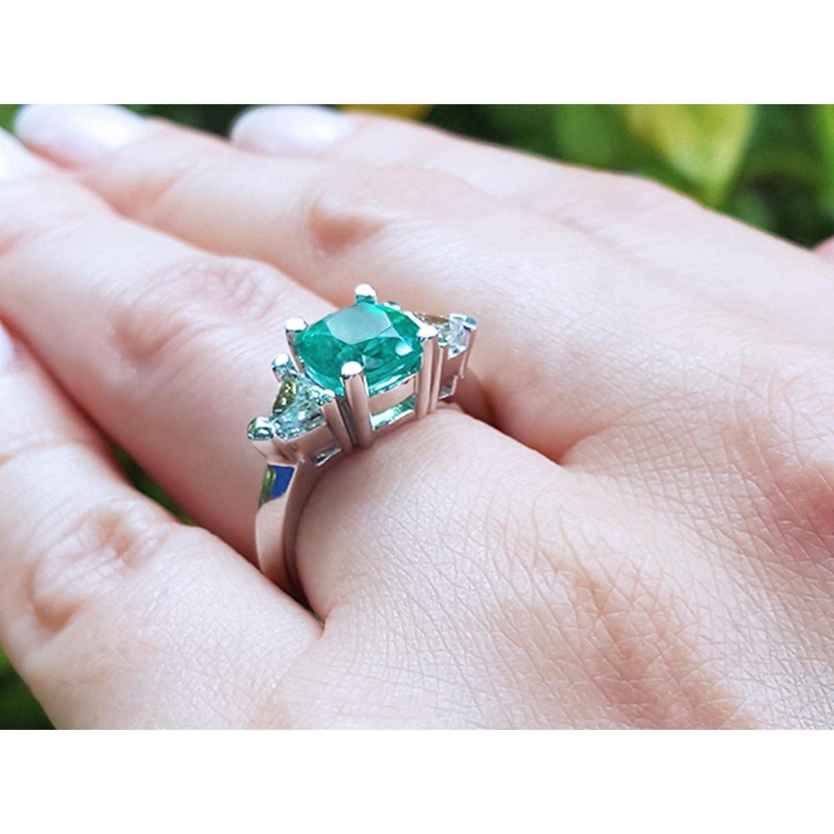 May's birthstone emerald ring cushion cut trillion side diamonds hand against a blurred natural background.