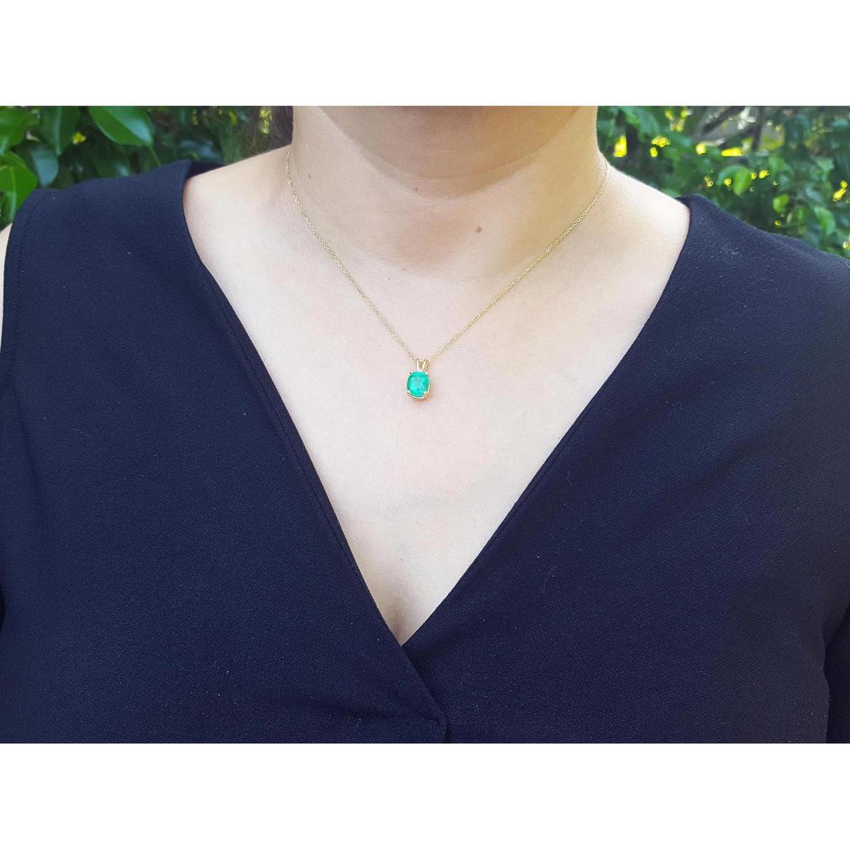 A woman wearing a necklace with a cushion emerald pendant against a blurred green background.