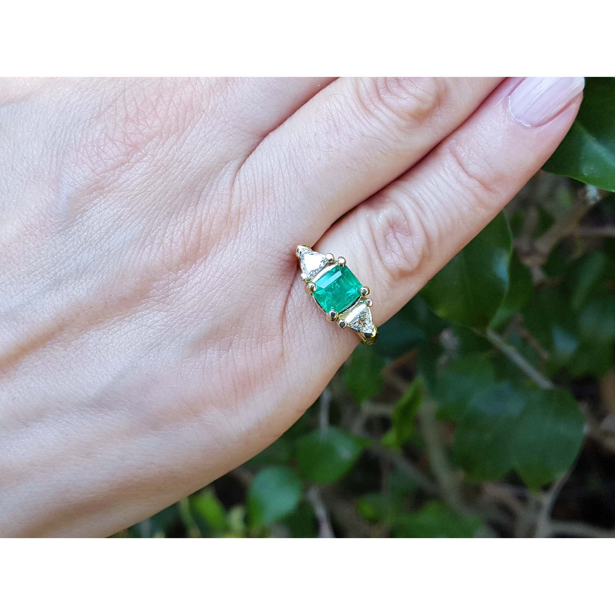 A 18k yellow gold emerald ring with trillion side diamonds on a hand against a natural background.